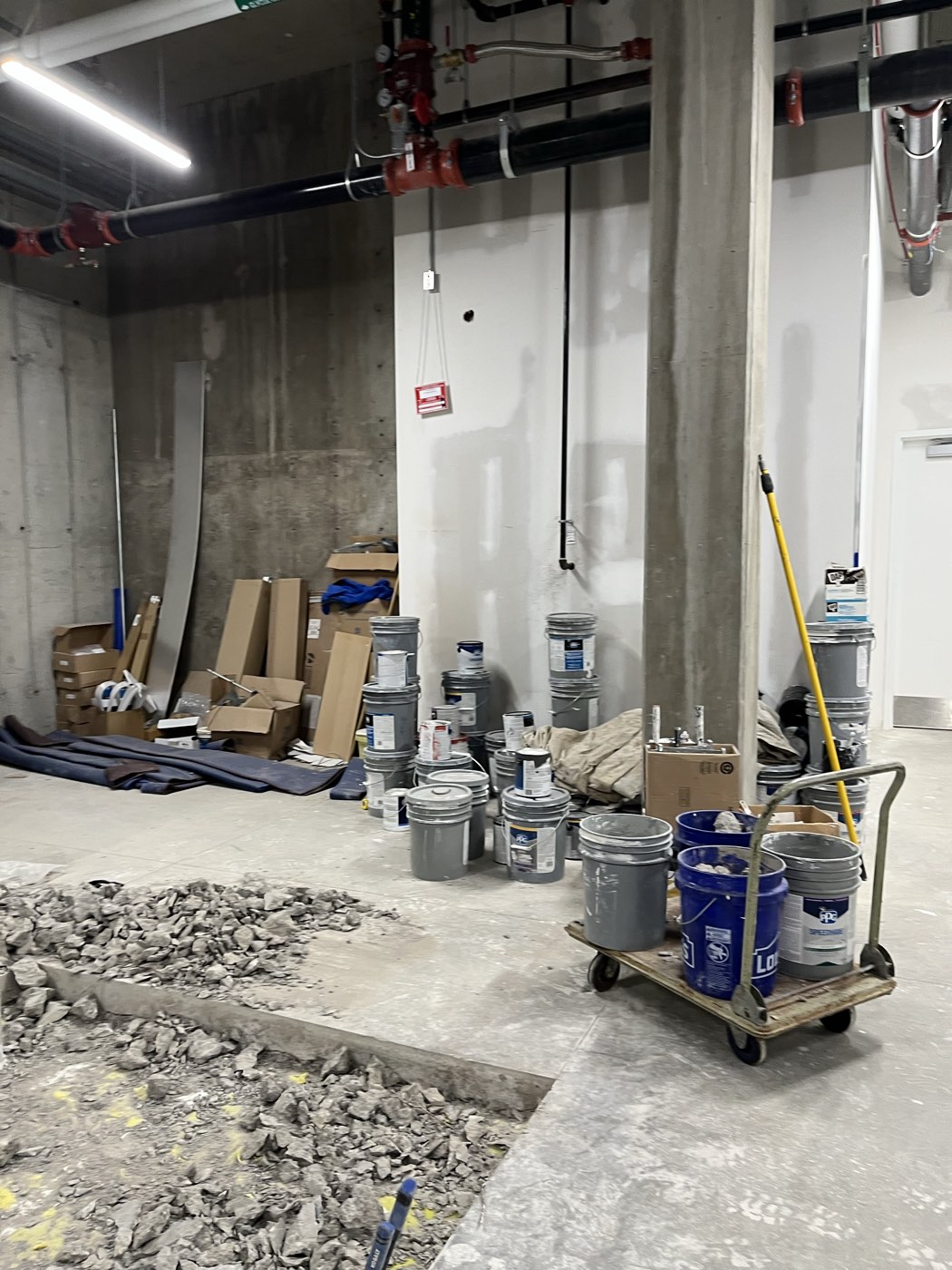 The future lab space before renovation: bare concrete walls and floor with a section broken out, stacked cardboard and paint buckets along one wall, and exposed pipework overhead.